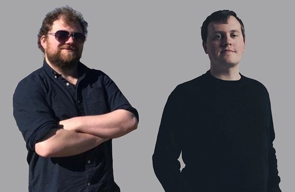 The performers standing against a gray background, facing the camera