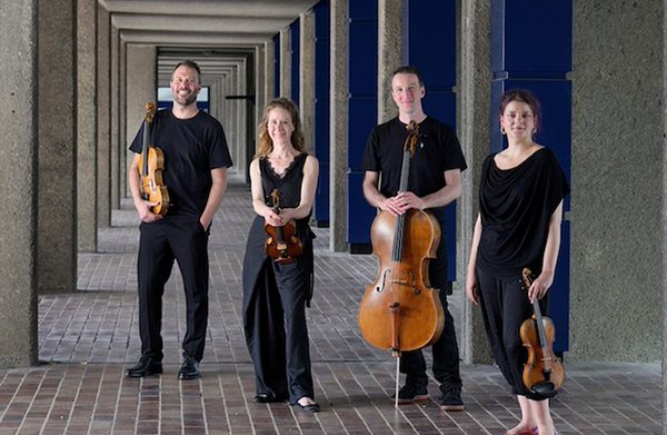 The performers standing on a tiled walkway, some of them holding instruments, and facing the camera