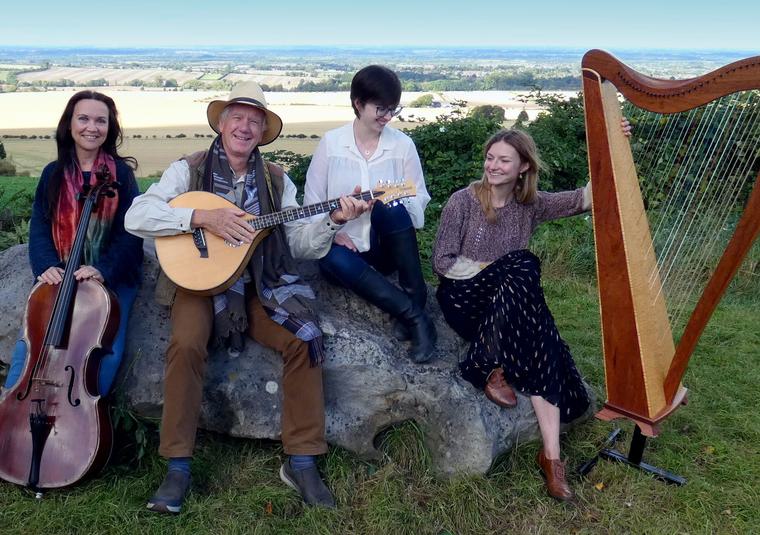 A group of four musicians holding a cello, mandolin and harp whilst sat on a rock in the countryside.
