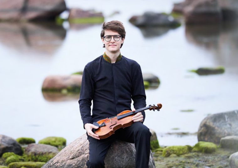 Performer sitting in front of a lake, holding an instrument, and looking at the camera