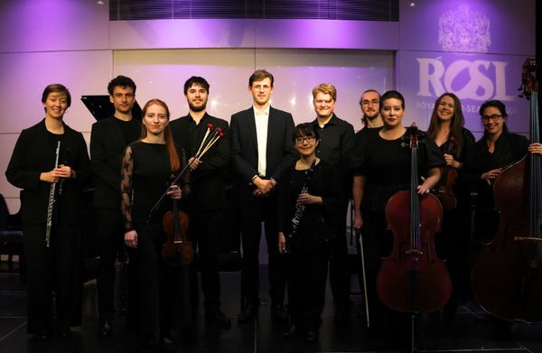 The performers, holding their instruments, and depicted in front of a white and purple backdrop