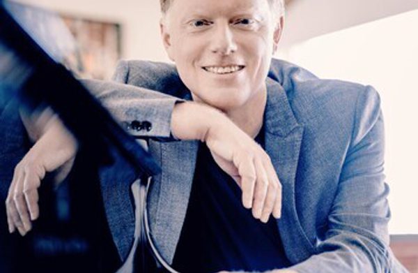 The performer sitting beside a piano looking at the camera