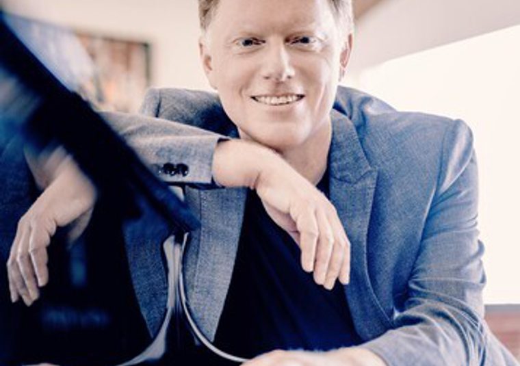 The performer sitting beside a piano looking at the camera