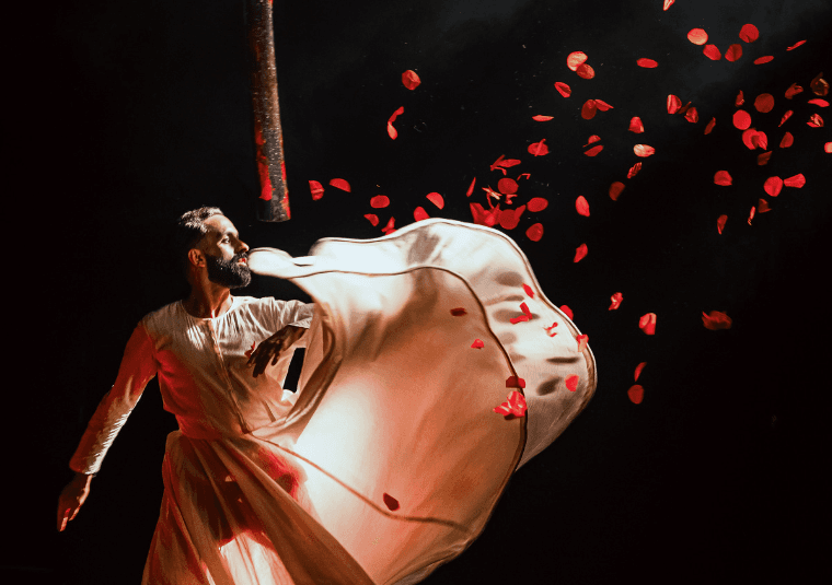 A man is stood wearing a white gown which is backlit. Part of the gown is suspended mid air and shadows can be seen through. Red petals are floating in the air against a dark backdrop.